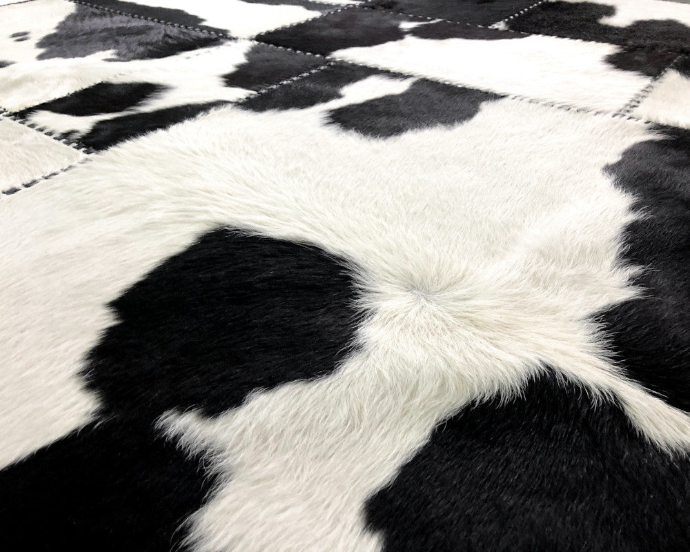 A square patchwork cowhide rug featuring a black and white cow print pattern made of stitched rectangular panels.