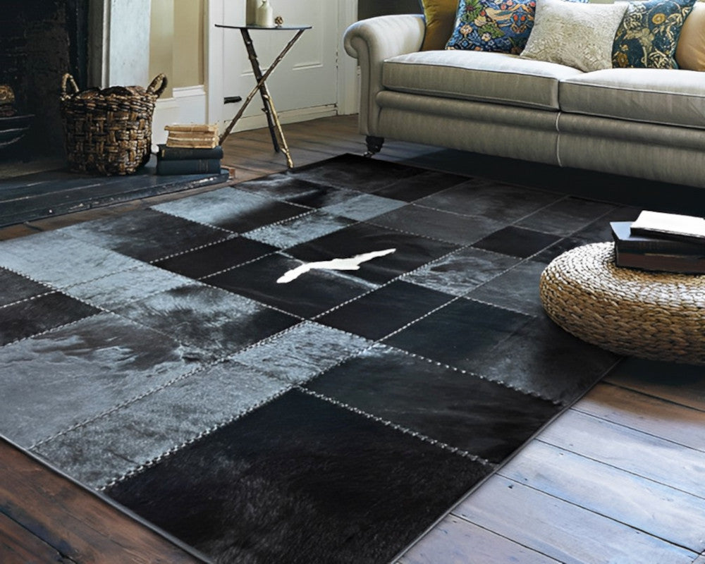 A large patchwork rug in black, charcoal, and gray tones with stitched square panels, shown in a styled living room setting with a sofa, side table, and wooden floor.
