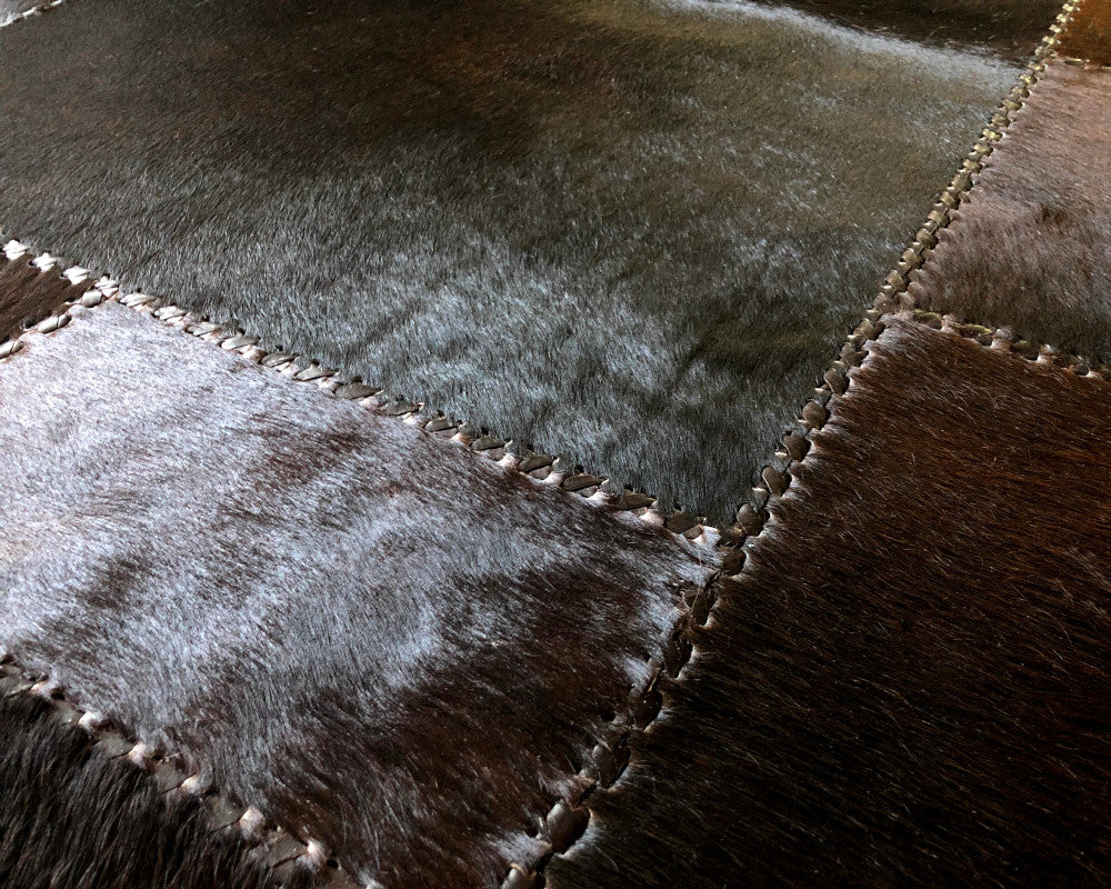 A square patchwork wall hanging or rug featuring rectangular sections of dark brown, black, and blue-gray textured leather or faux leather, stitched together with visible white stitching lines.