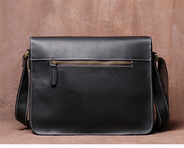 A black leather crossbody bag with white contrast stitching and a gold zipper on the front flap, shown against a textured brown background.