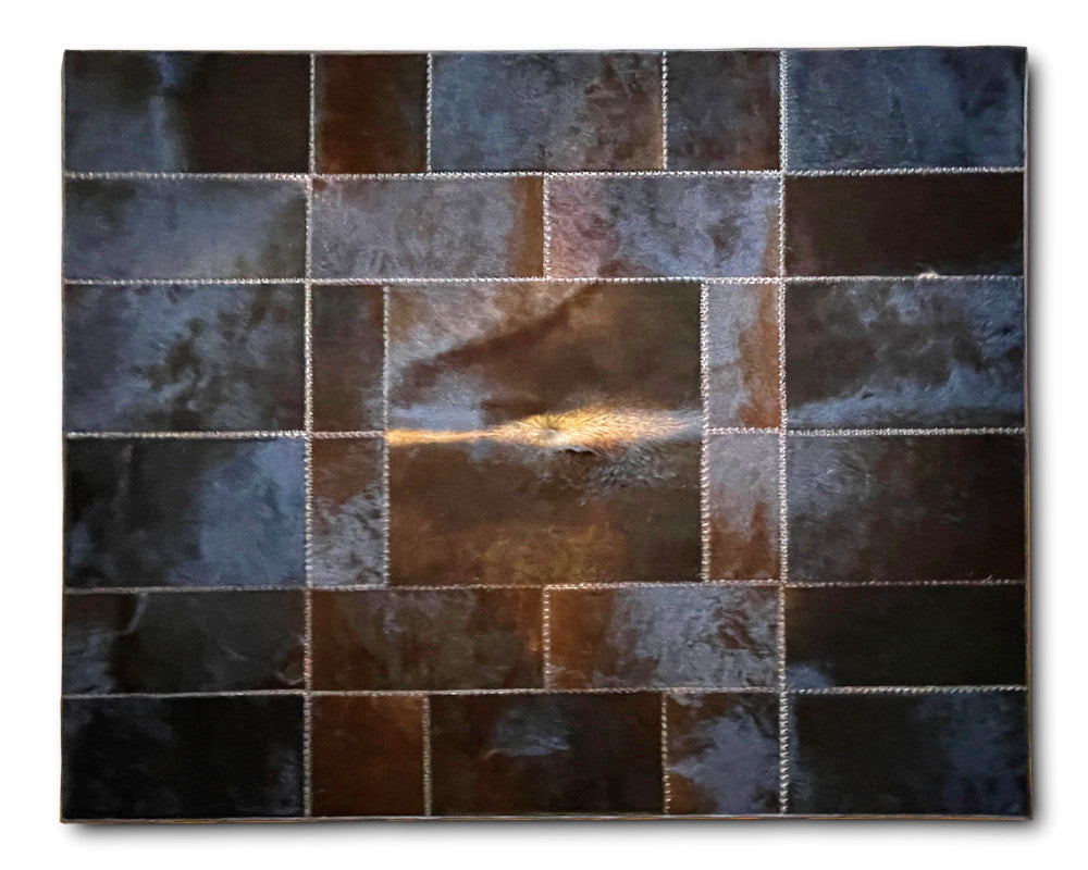 A rectangular patchwork leather rug featuring a grid pattern of stitched square panels in dark brown, bronze, and gray tones, with a small warm light reflection in the center, shown against a plain white background.