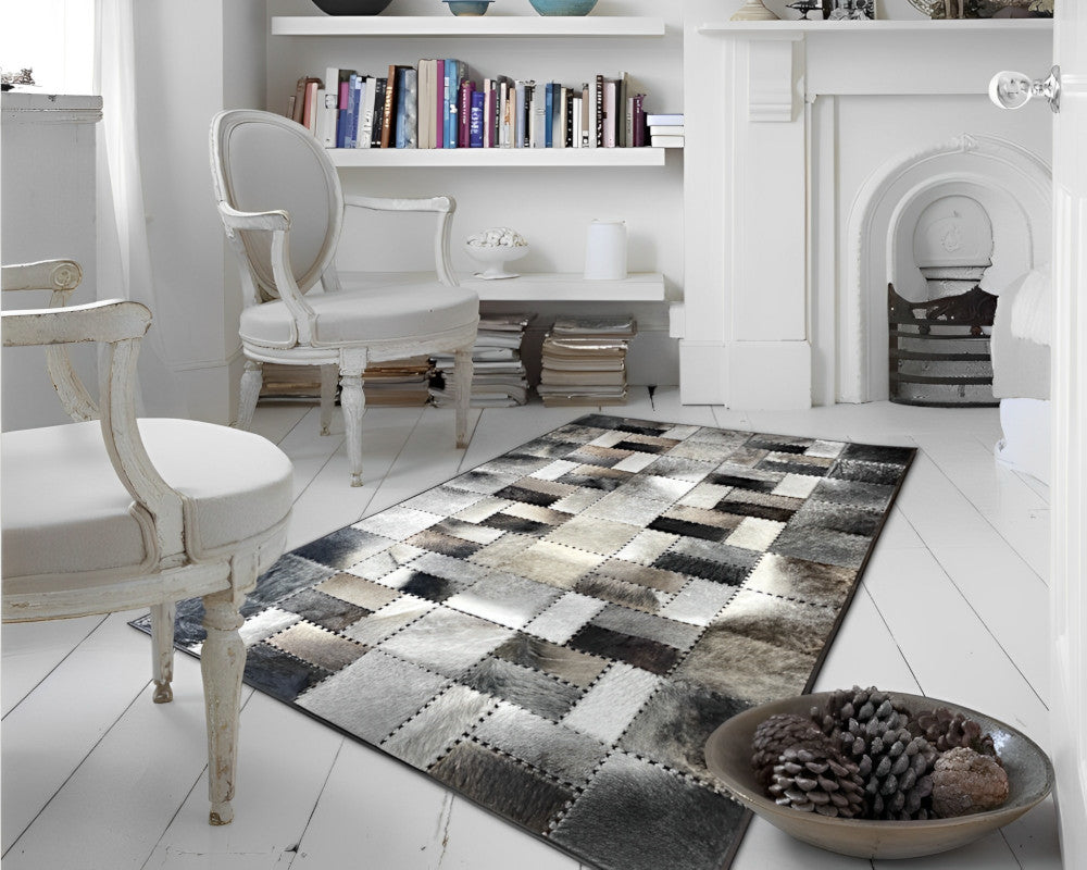 A rectangular patchwork rug with a cowhide-inspired pattern in black, gray, beige, and white, styled on a white wooden floor in a bright, classic living room.