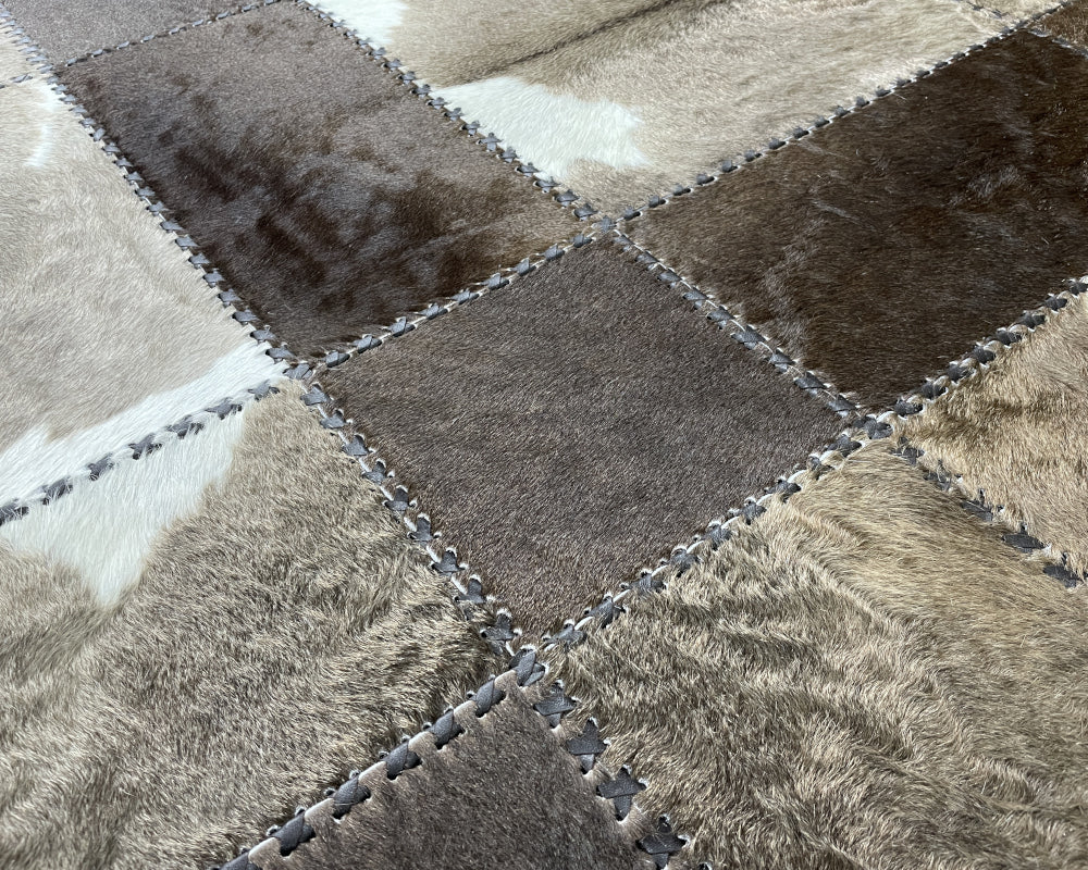 A square patchwork rug made of irregular cowhide-style panels in mixed gray, taupe, brown, and off-white tones, stitched together with a dark border.