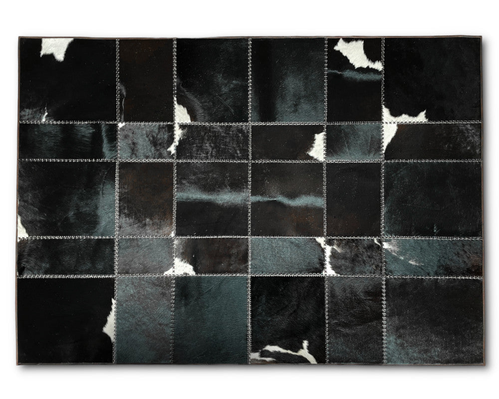 A rectangular patchwork rug made of stitched square panels in black, charcoal, and dark gray with some white hide-like patches, shown flat against a light gray background.