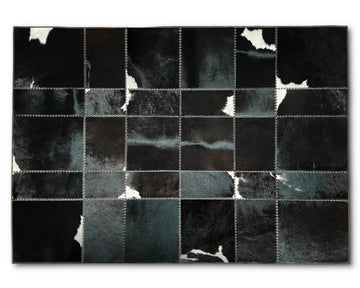 A rectangular patchwork rug made of stitched square panels in black, charcoal, and dark gray with some white hide-like patches, shown flat against a light gray background.