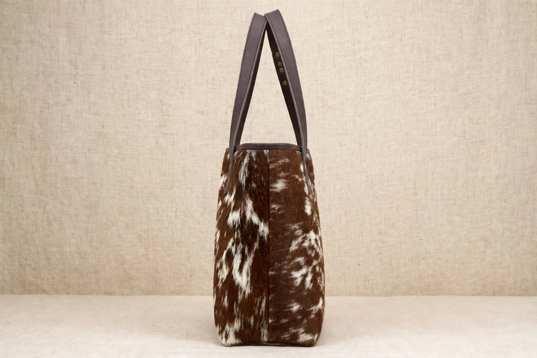Genuine leather cowhide tote bag with natural hair-on-hide texture, leather shoulder handles, stitched detailing, and brass-tone rivets