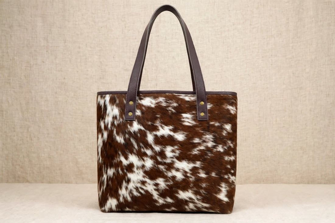 Genuine leather cowhide tote bag with natural hair-on-hide texture, leather shoulder handles, stitched detailing, and brass-tone rivets